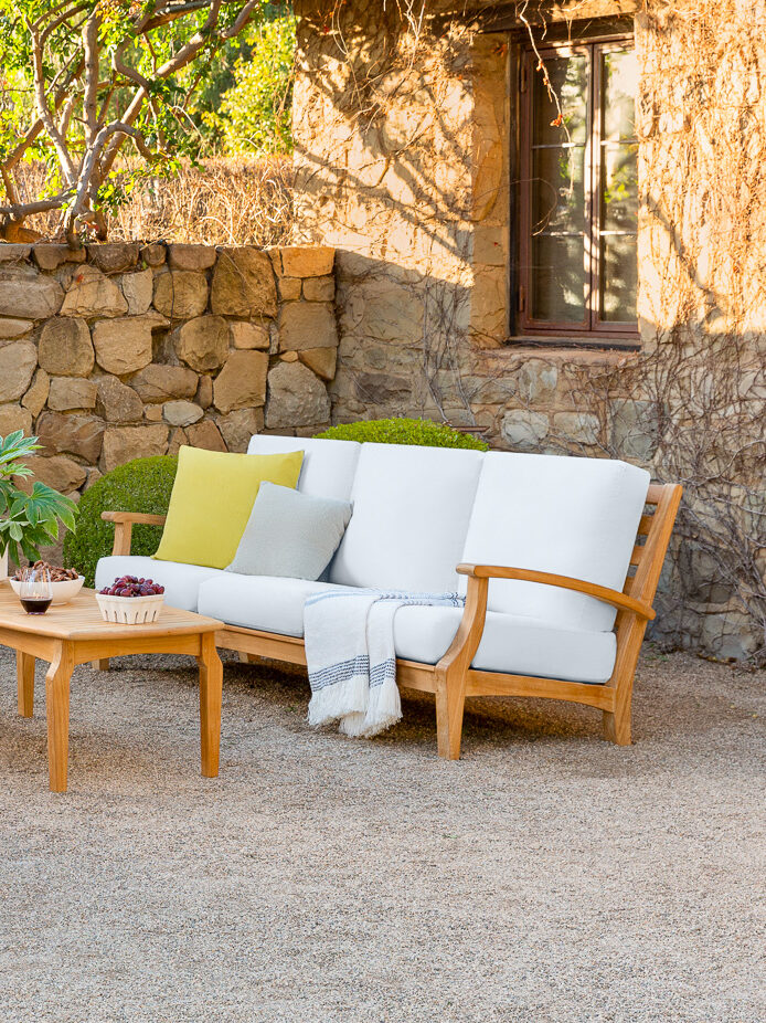Outdoor patio area with wooden furniture featuring white cushions, a stone fireplace, a coffee table with decor, and various plants in a sunlit garden setting.