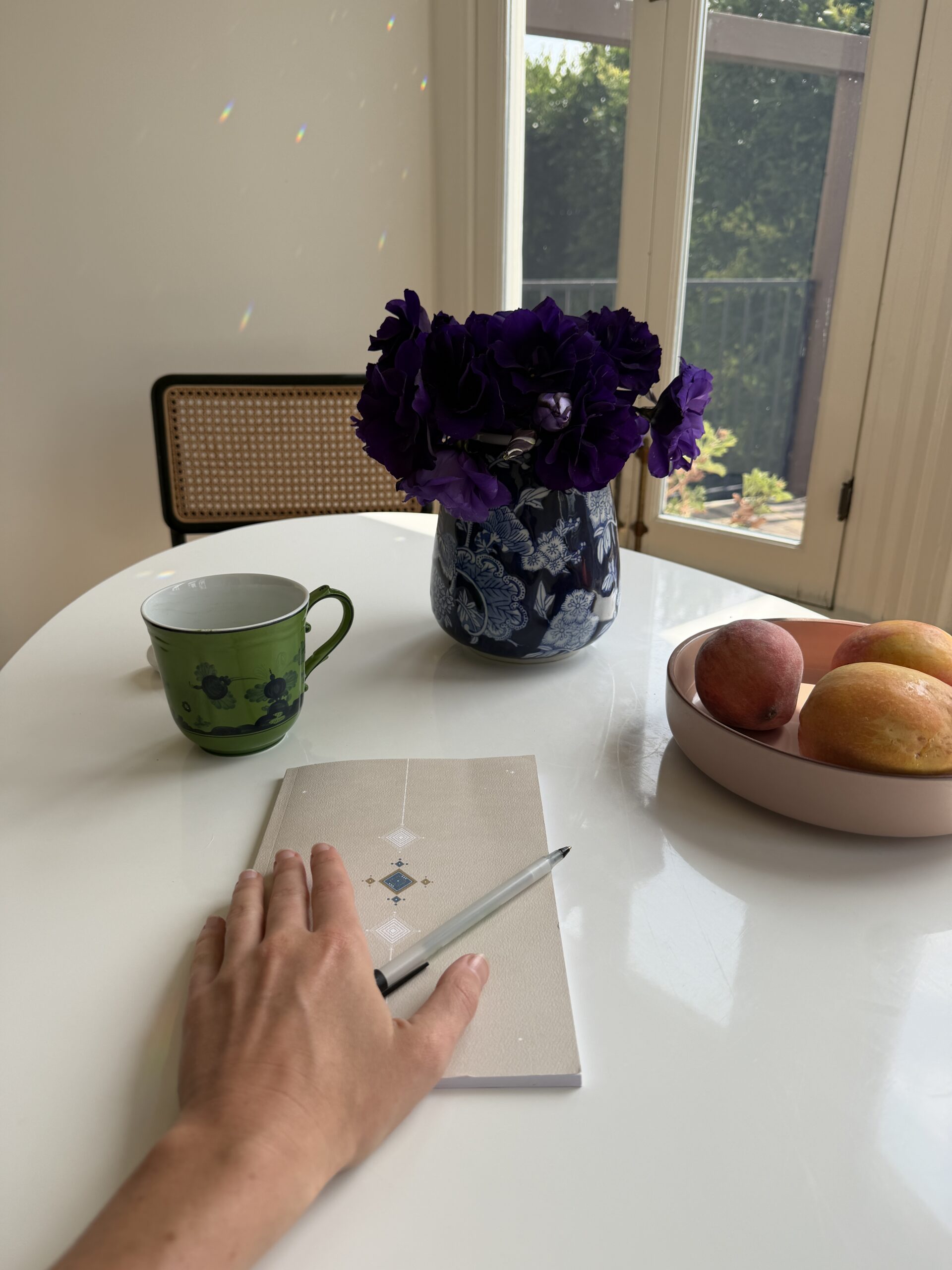 A hand rests on a closed notebook with a pen on a white table, next to a green mug, a vase of purple flowers, and a bowl of peaches and a mango.