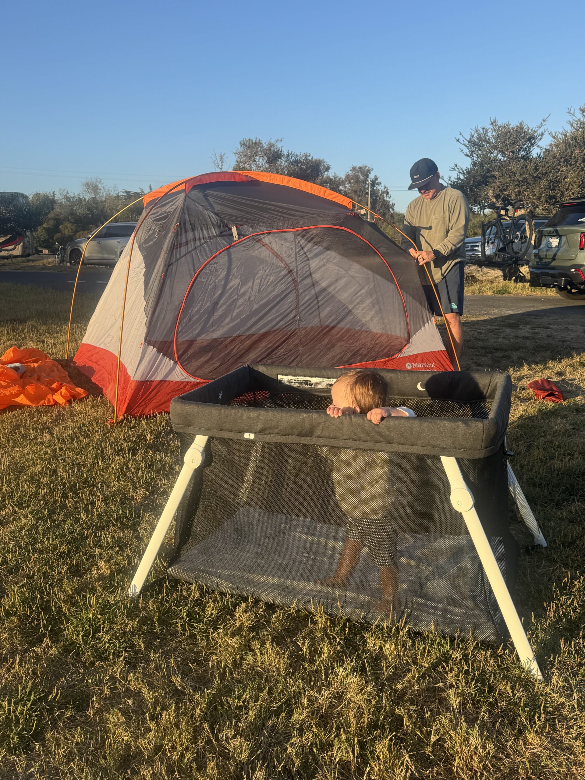 A man sets up a red and gray tent on grass while a toddler stands in a black playpen outdoors.