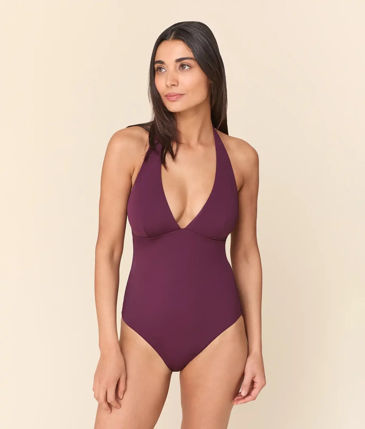 Woman wearing a maroon one-piece halter swimsuit stands against a plain beige background, looking off to the side.
