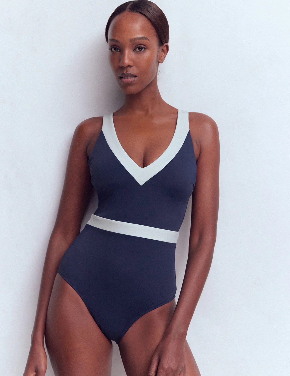 A woman stands against a plain light background wearing a navy blue one-piece swimsuit with white trim along the neckline and waist.