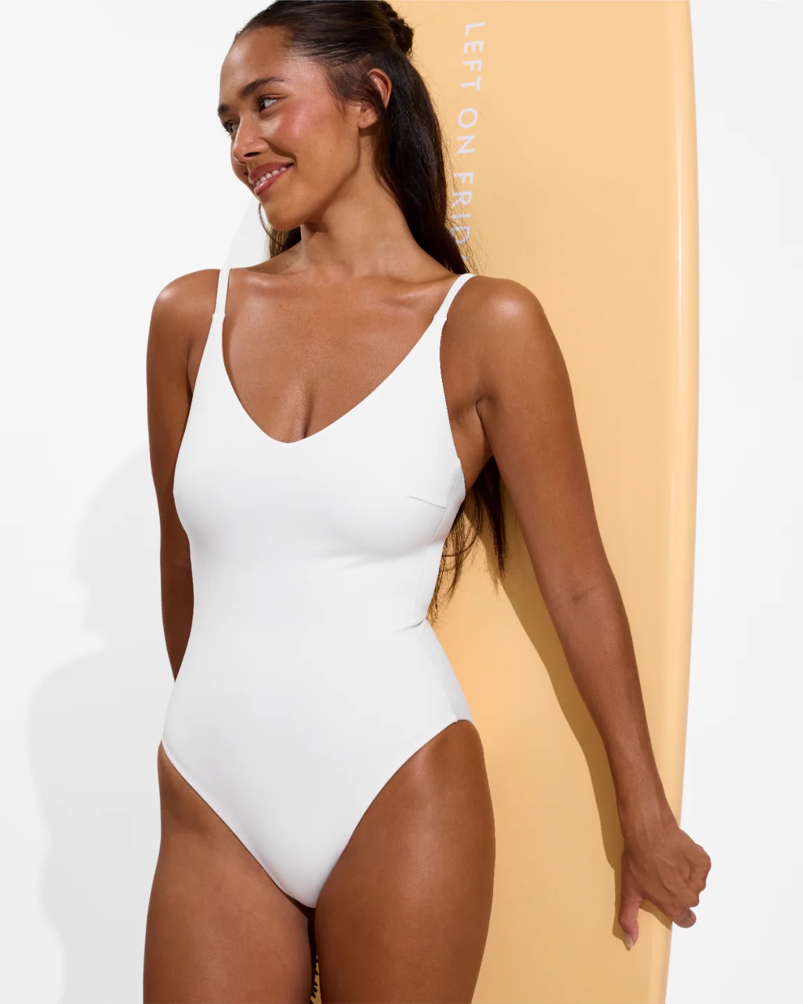 Woman in a white one-piece swimsuit stands and smiles next to a yellow surfboard against a plain background.