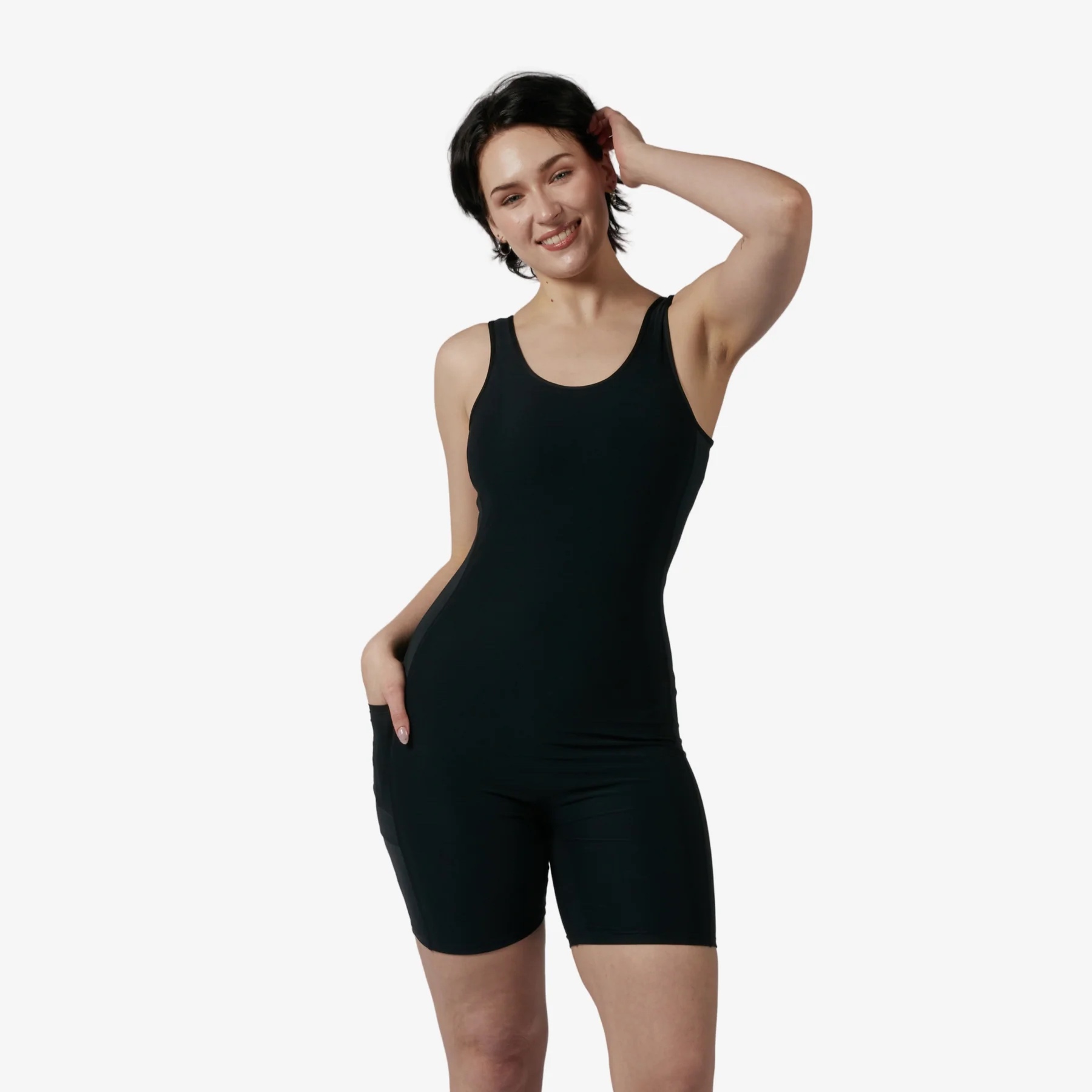 A person stands against a white background wearing a black sleeveless fitted romper, smiling with one hand on their head and the other in a pocket.