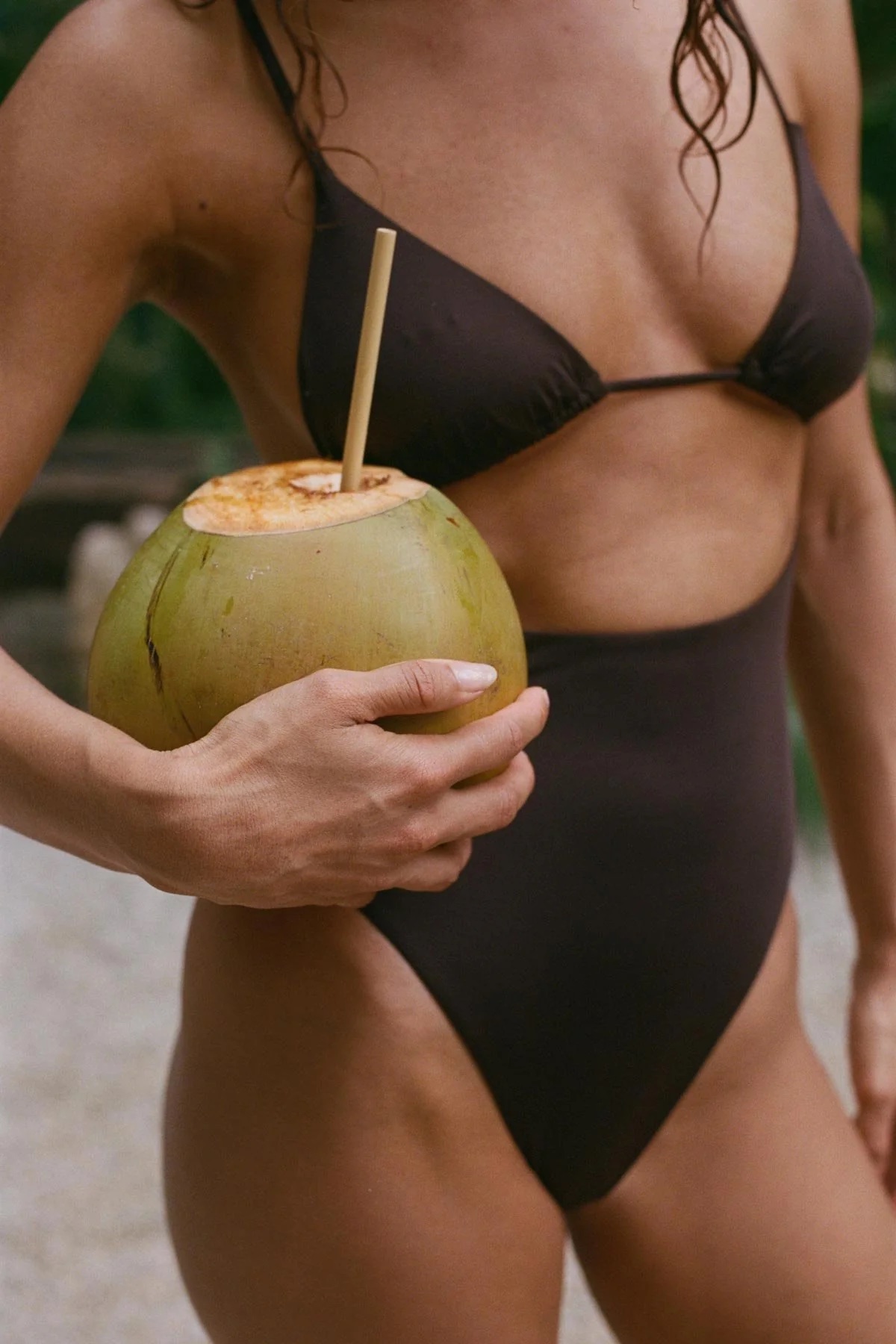A person in a dark bikini holds a green coconut with a straw inserted into it.