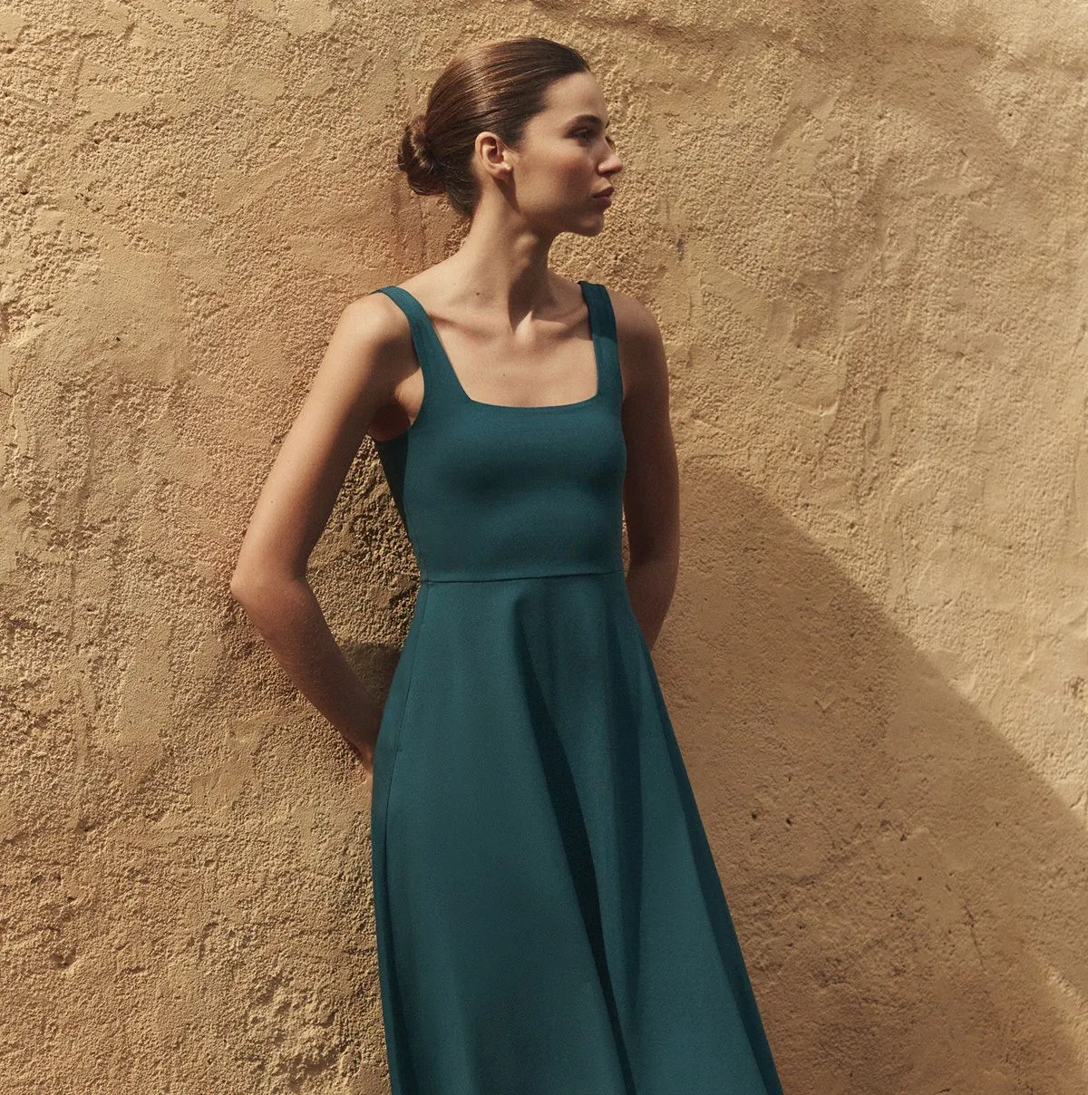 A woman in a teal sleeveless dress stands against a textured beige wall, looking to her left with her hair pulled back in a neat bun.