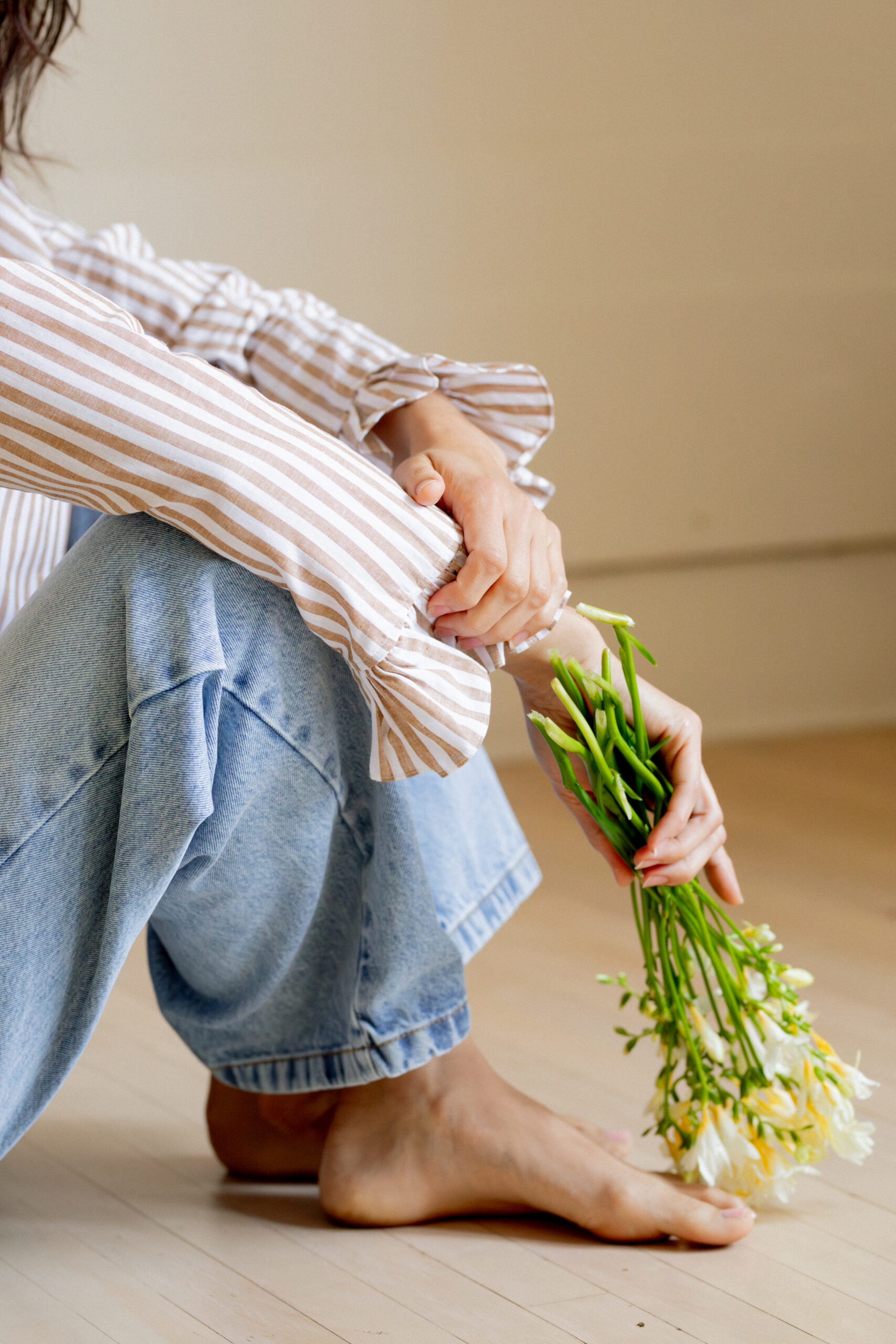 A person in a striped shirt and jeans sits barefoot on a wooden floor, holding a small bouquet of wilted yellow flowers.