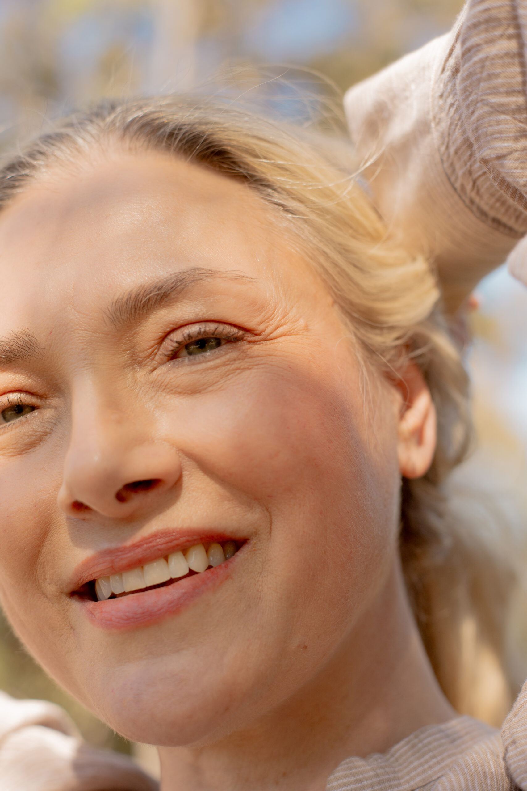 Close-up of a woman with light skin and blonde hair smiling outdoors, with sunlight and shadows on her face.