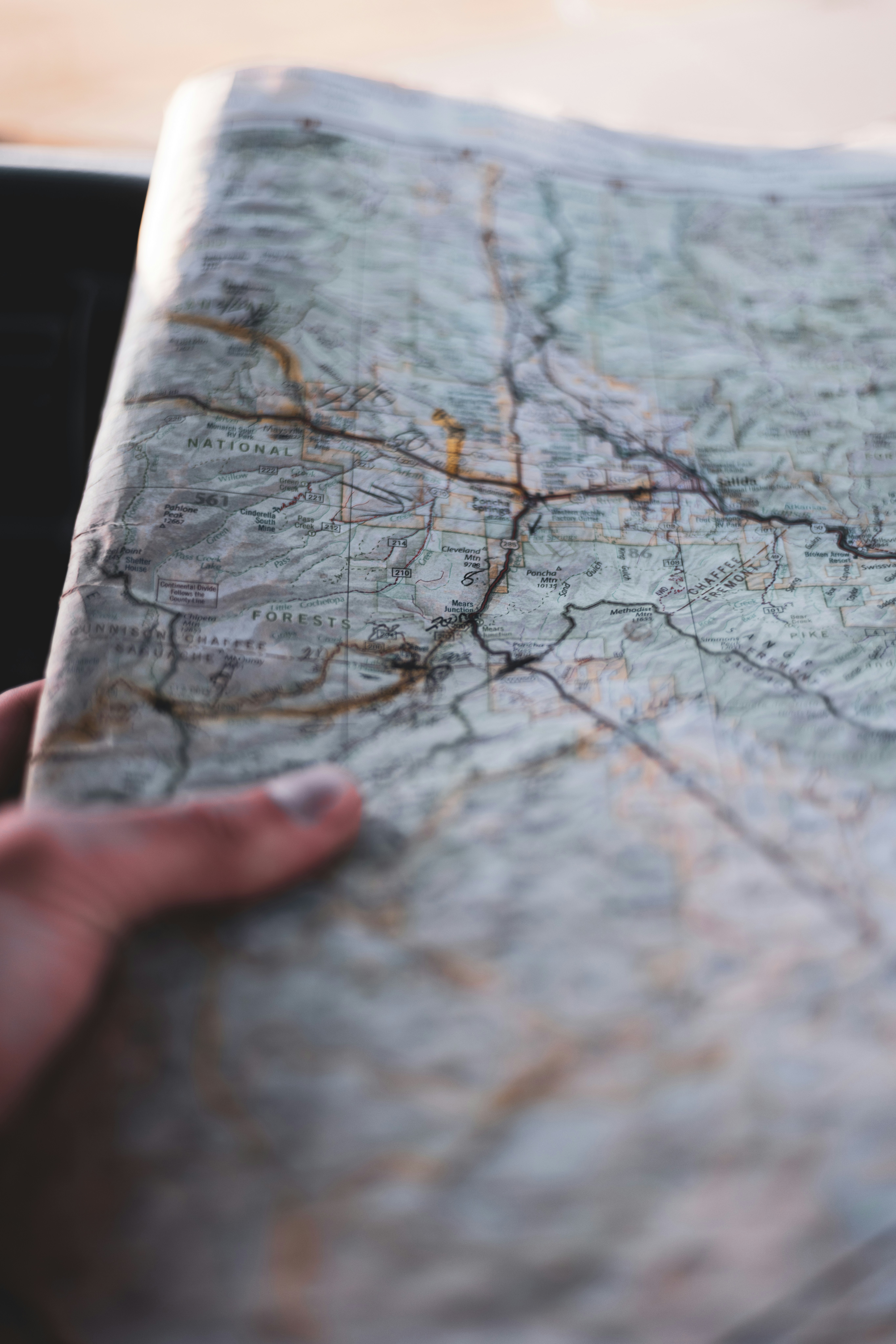 A hand holds a folded paper map showing detailed topography, roads, and place names in a natural landscape area.