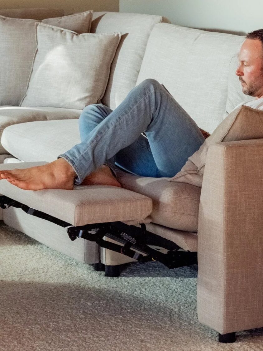 Man sits and relaxes on a light-colored sectional sofa with a recliner extended, next to a round wooden coffee table and a side table with stacked books.