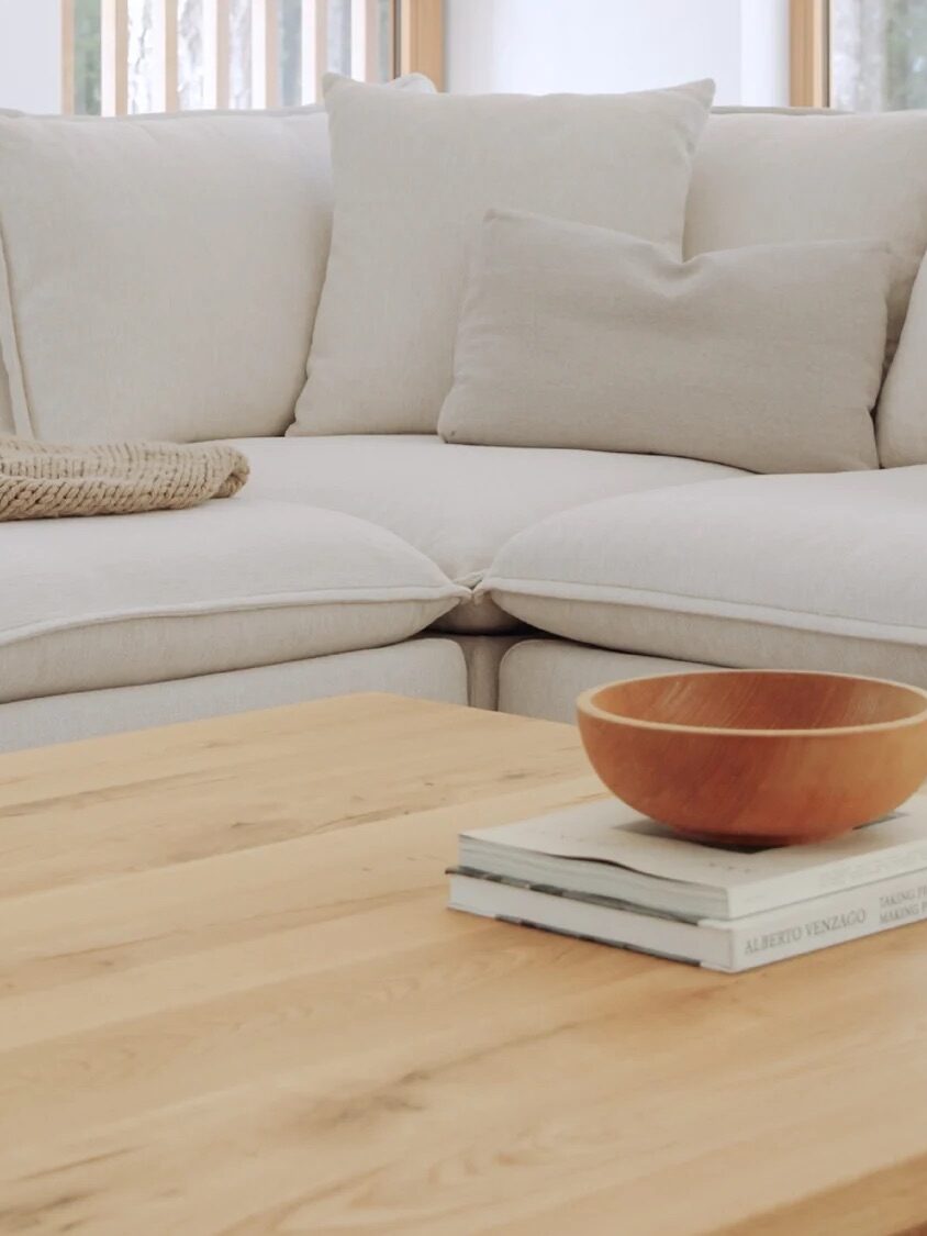 A light-colored sectional sofa with a blanket and a small dog on it sits behind a wooden coffee table with books and a bowl in a bright, modern living room.