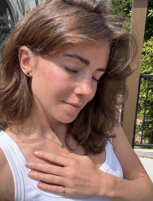 A woman with brown hair closes her eyes and places her hand on her chest while standing outdoors in sunlight, wearing a white tank top and a necklace.