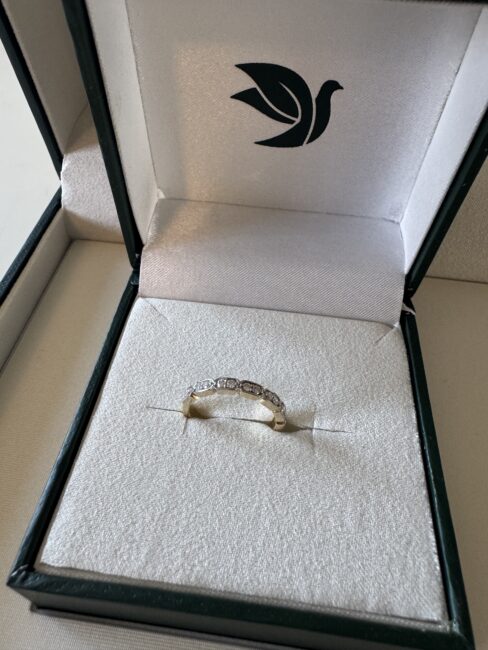 A gold ring with several small diamonds is displayed in a white cushioned jewelry box with a green bird logo on the inside lid.