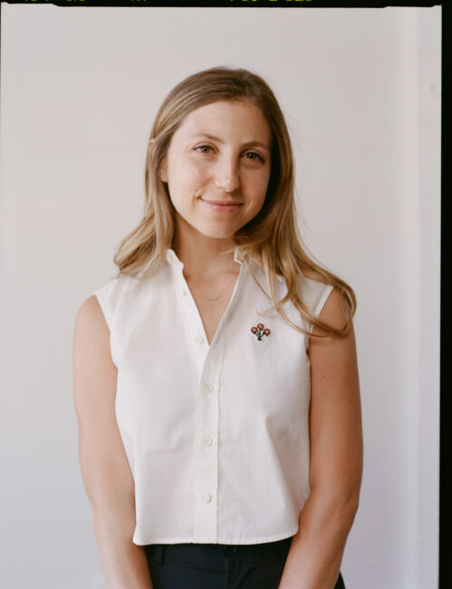 A woman with long hair wears a sleeveless white button-up shirt with a floral pin, standing against a plain light background and smiling at the camera.