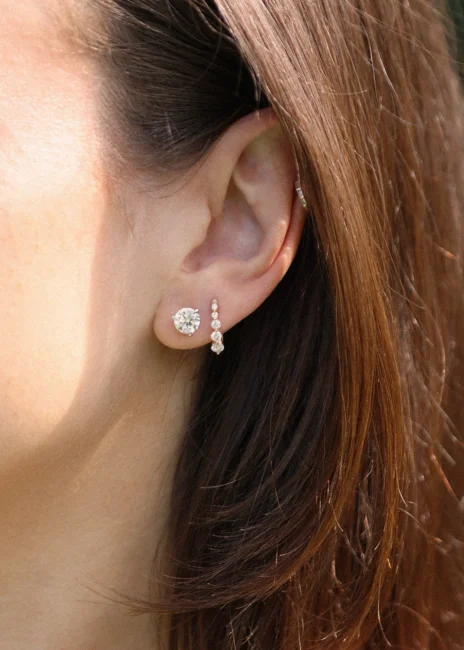 A close-up of a woman’s ear with two earrings: a large round stud and a vertical arrangement of small stones. Brown hair partially covers the ear.