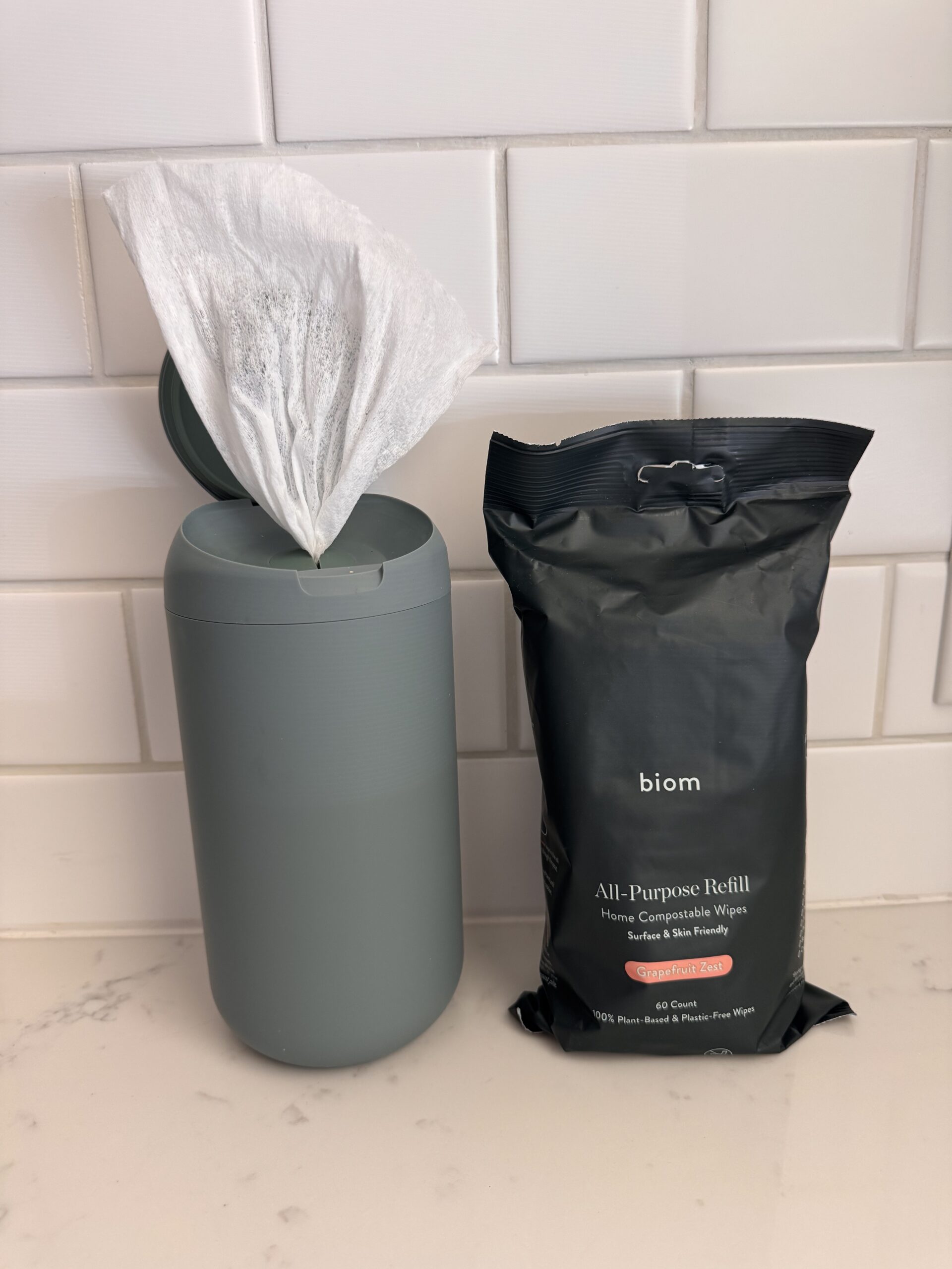A gray canister with a wipe sticking out stands next to a black refill pack labeled "biom all-purpose refill," both on a white tiled countertop.