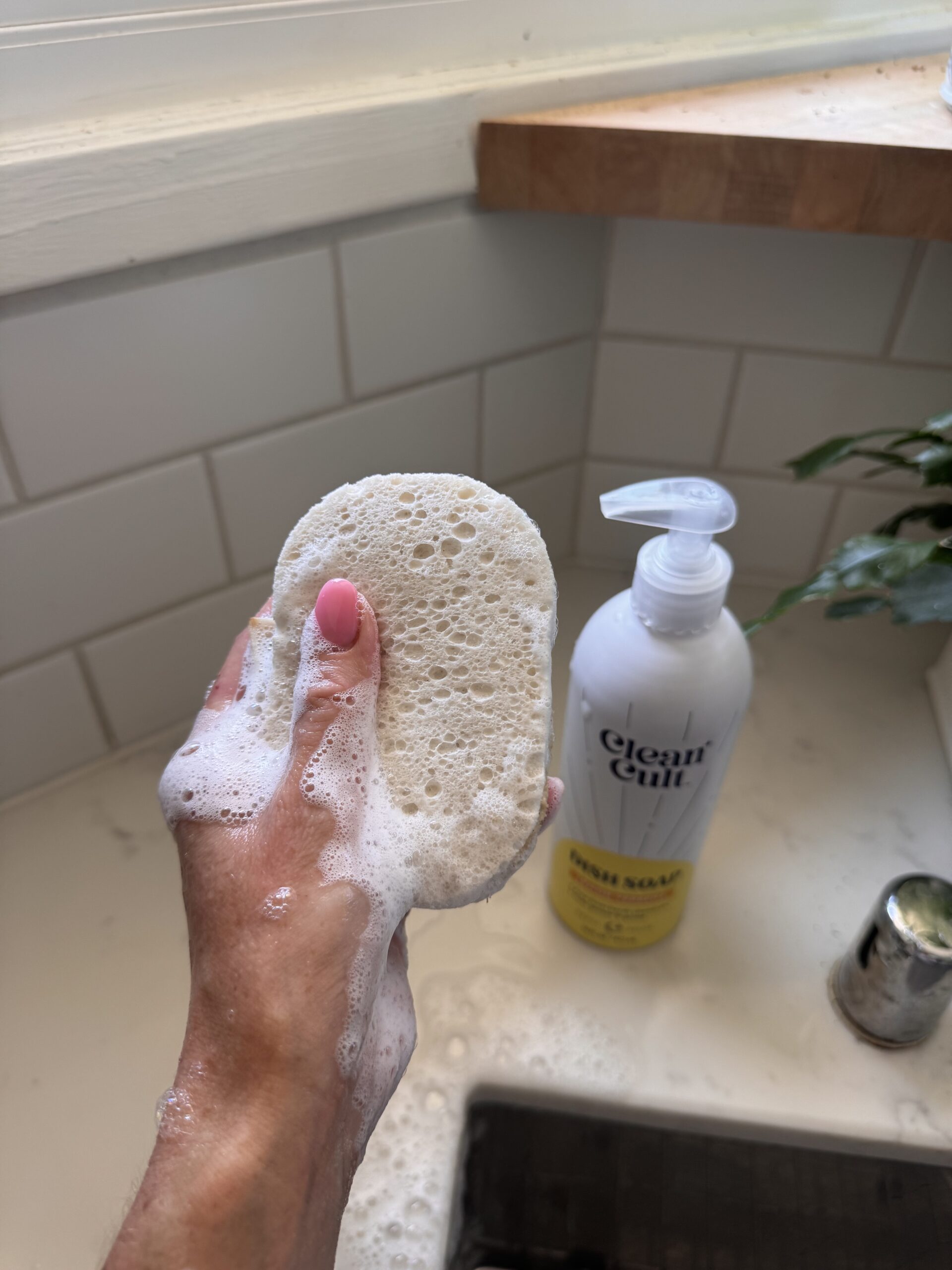 A hand with pink nail polish holds a soapy sponge near a bottle of liquid soap on a bathroom sink.