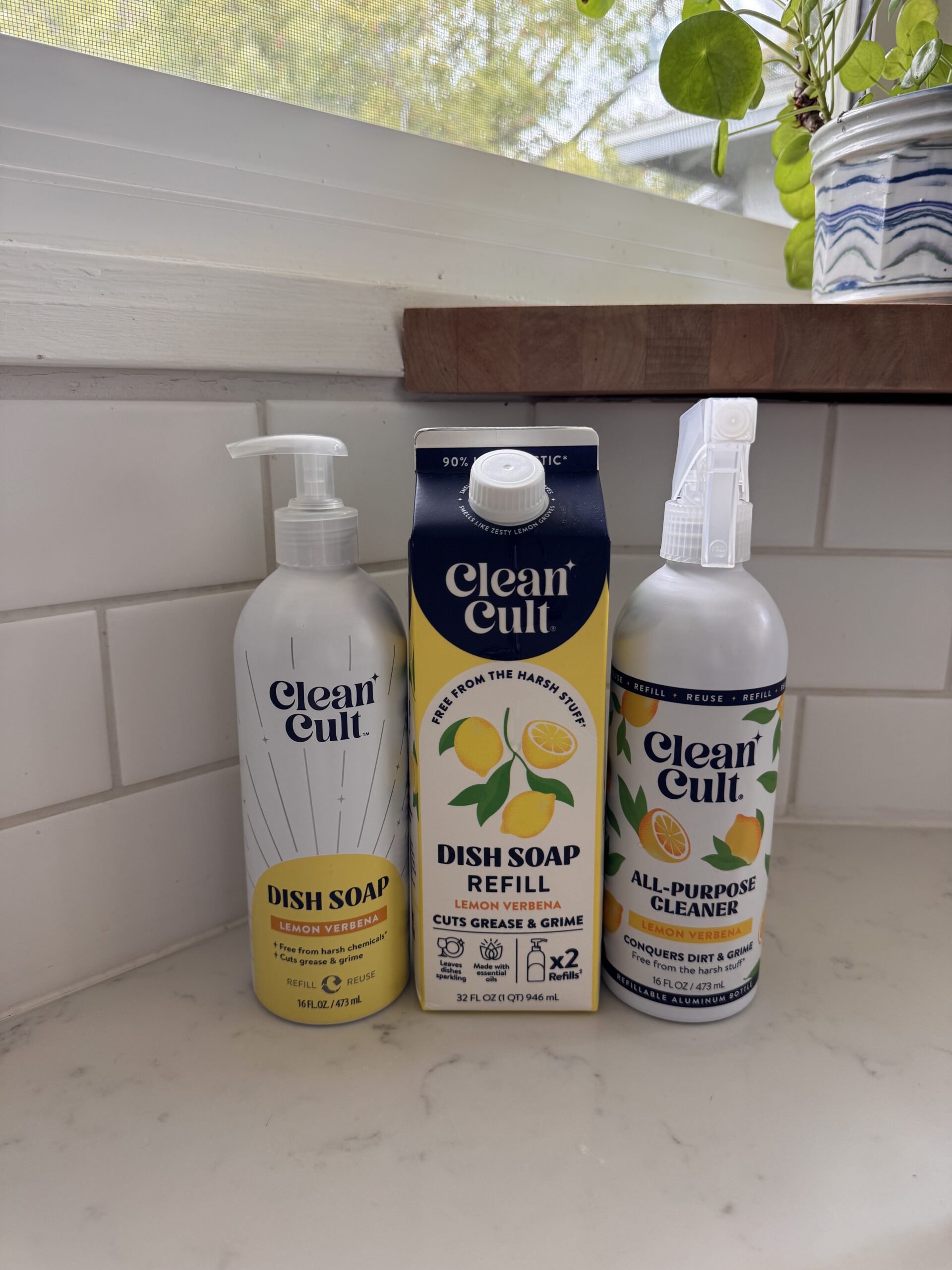 Three Clean Cult products—dish soap, dish soap refill, and all-purpose cleaner—are arranged on a kitchen countertop near a window with a potted plant.