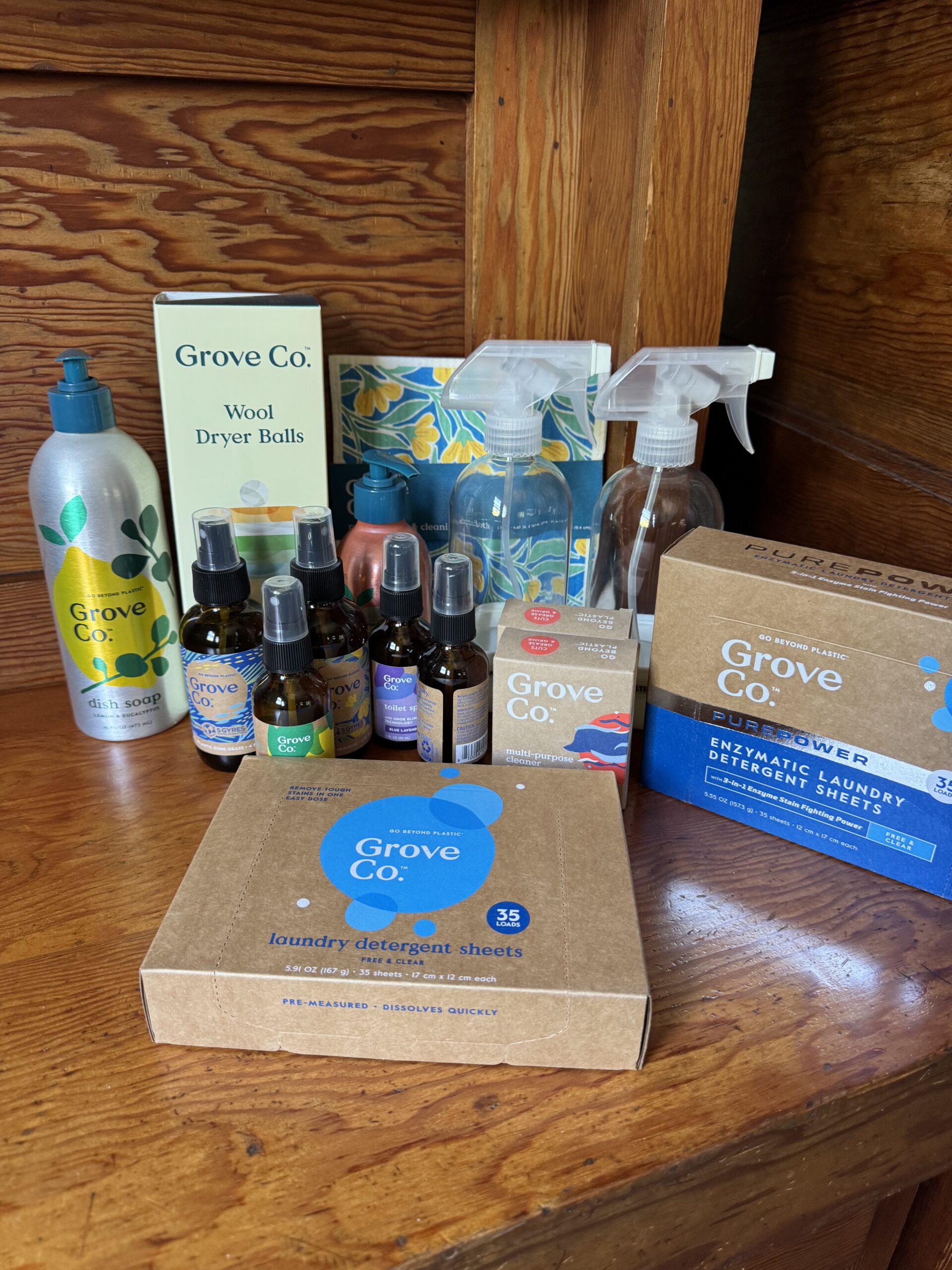Grove Co. cleaning and household products, including detergent sheets, sprays, wool dryer balls, and essential oil bottles, arranged on a wooden surface.