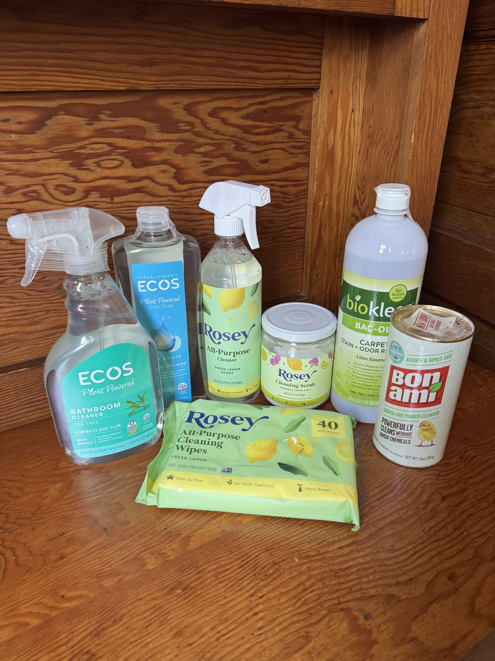 Seven eco-friendly cleaning products, including sprays, wipes, and powders, are arranged on a wooden surface in front of a wooden wall.
