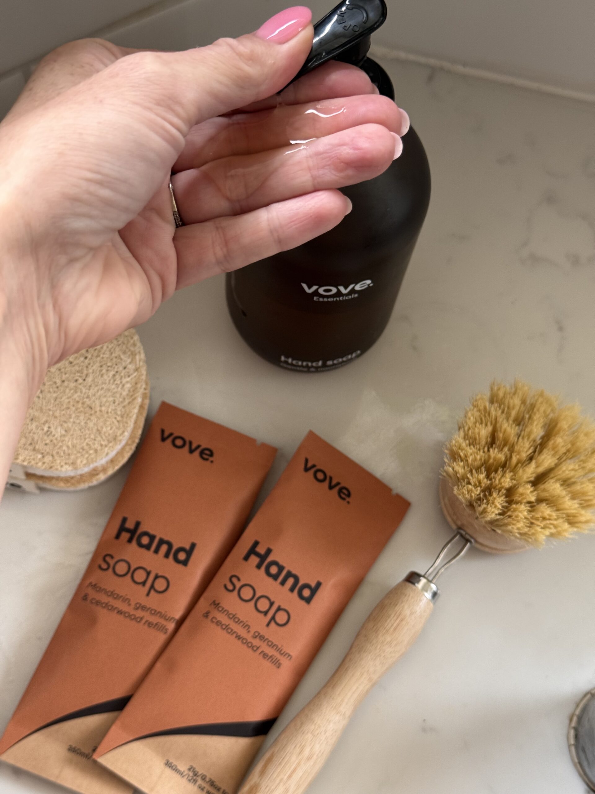 A hand dispenses liquid from a Vove hand soap bottle, with two orange soap refill packets, a scrub brush, and a round sponge on a white countertop.
