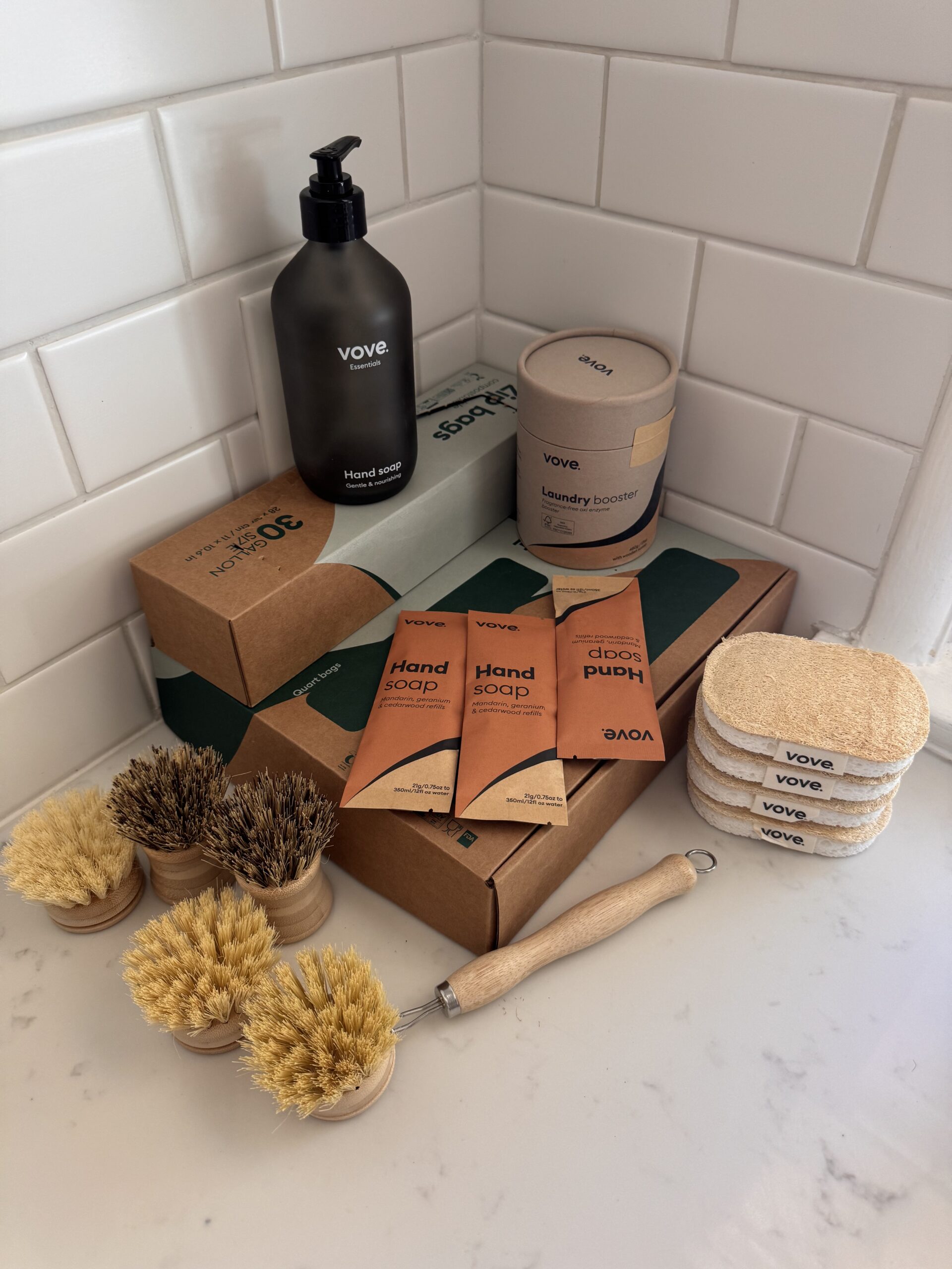 A collection of eco-friendly cleaning products, including brush sets, sponges, soap packets, laundry booster, and boxed soap, arranged neatly on a white bathroom counter.